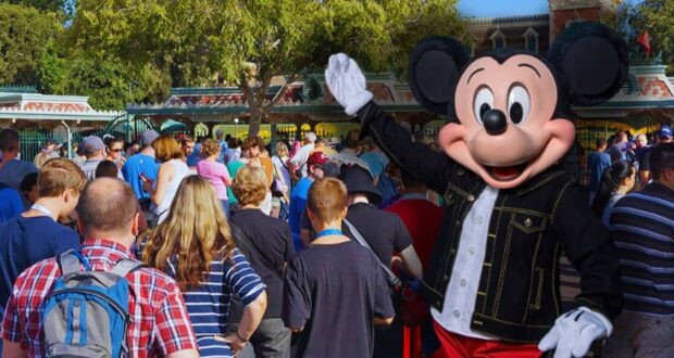 Mickey Mouse waves to a large crowd of people at an amusement park entrance. The crowd consists of adults and children, some wearing backpacks and hats, surrounded by trees and park buildings in the background.