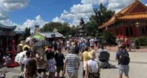 A large crowd of people walk down a wide path in a park or festival setting. Traditional architectural structures with ornate roofs line the walkway, and the sky is partly cloudy.