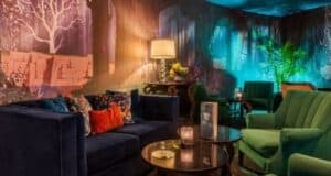 A cozy room with dark blue and green velvety chairs and a matching sofa. Ambient lighting highlights a mural of a mystical forest. A table holds a menu and candles, and a floor lamp adds a warm glow to the setting.