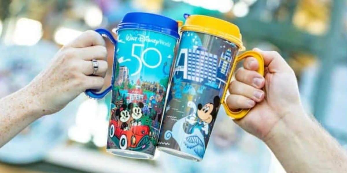 Two hands holding colorful Disney-themed mugs, each adorned with images of Mickey Mouse and Disney landmarks, against a blurred background. The mugs bear "Walt Disney World 50" in celebration of the park's anniversary.