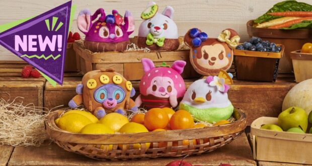 A variety of fruit-themed plush toys are displayed on wooden crates surrounded by baskets of colorful fruits and vegetables. A purple label with the word "NEW!" is on the left side.