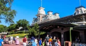People walk toward the entrance of a theme park on a sunny day. The building features a clock tower and intricate architectural details, with a well-maintained garden in front. Trees and a clear blue sky are in the background.
