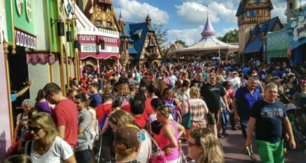 A large crowd of people walking through a busy amusement park area with brightly colored buildings and a carousel in the background. The sky is clear and blue with a few clouds. The scene is lively and bustling with families and visitors.