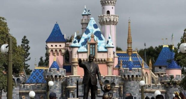 A statue of a man holding hands with a mouse character stands prominently in front of a whimsical castle with blue and pink spires. Visitors gather around, enjoying the vibrant and colorful scene. Trees and lamps frame the scene in an outdoor setting.