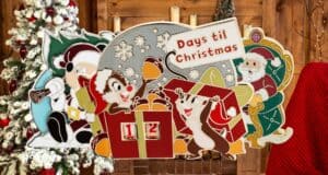 Christmas countdown sign with cartoon reindeer and elves, set against a festive wooden background. The sign reads "Days 'til Christmas" and has numbered blocks displaying "12." Decorated Christmas tree and red plaid chair in the background.