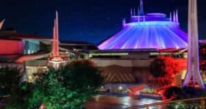 A nighttime view of a futuristic building with a domed roof illuminated in colorful lights, surrounded by trees and a clear, starry sky. The scene has vibrant red, blue, and purple hues.