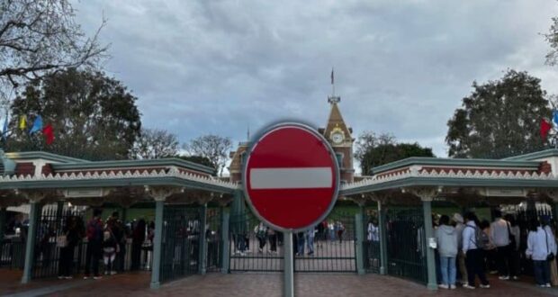 A "do not enter" sign in front of a theme park entrance, with a crowd of people gathered outside. The park features decorative gates and a clock tower, with trees and a cloudy sky in the background.