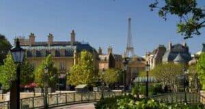 Immerse yourself in a picturesque European-style village, reminiscent of Epcot's charm, with pastel-colored buildings and grey rooftops. The Eiffel Tower graces the clear blue sky, while lush green trees and vintage street lamps complete this enchanting scene.