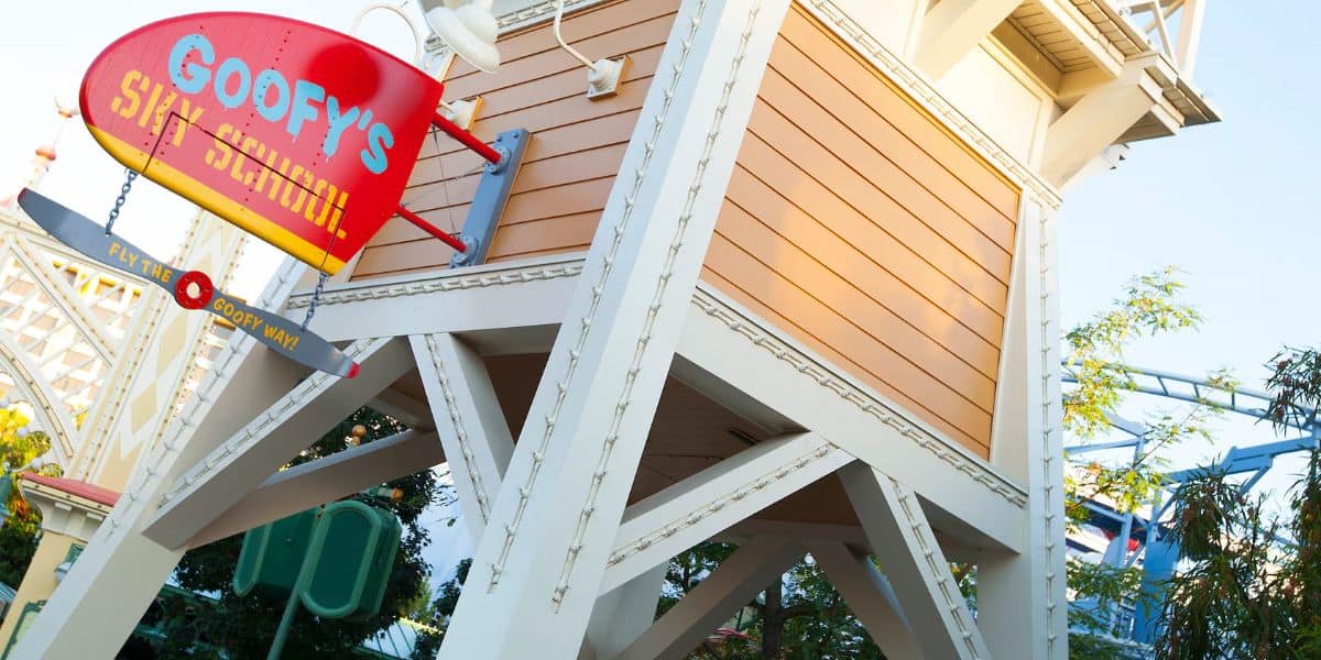 A close-up of a wooden tower structure with a red and yellow sign reading "Goofy's Sky School." The sign is decorated with an airplane motif. The building has white beams and orange paneling, surrounded by trees and a clear sky.