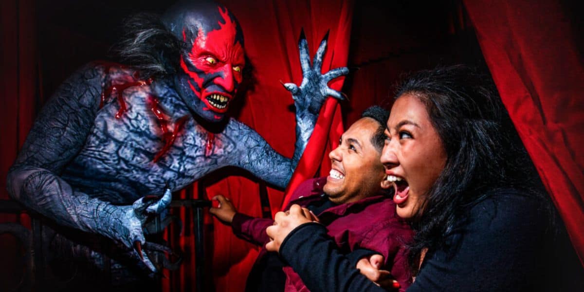 A couple sits in a haunted house attraction, looking terrified as a scary creature with red and black skin, sharp teeth, and long claws reaches out menacingly toward them. Red curtains and dark shadows enhance the eerie atmosphere.