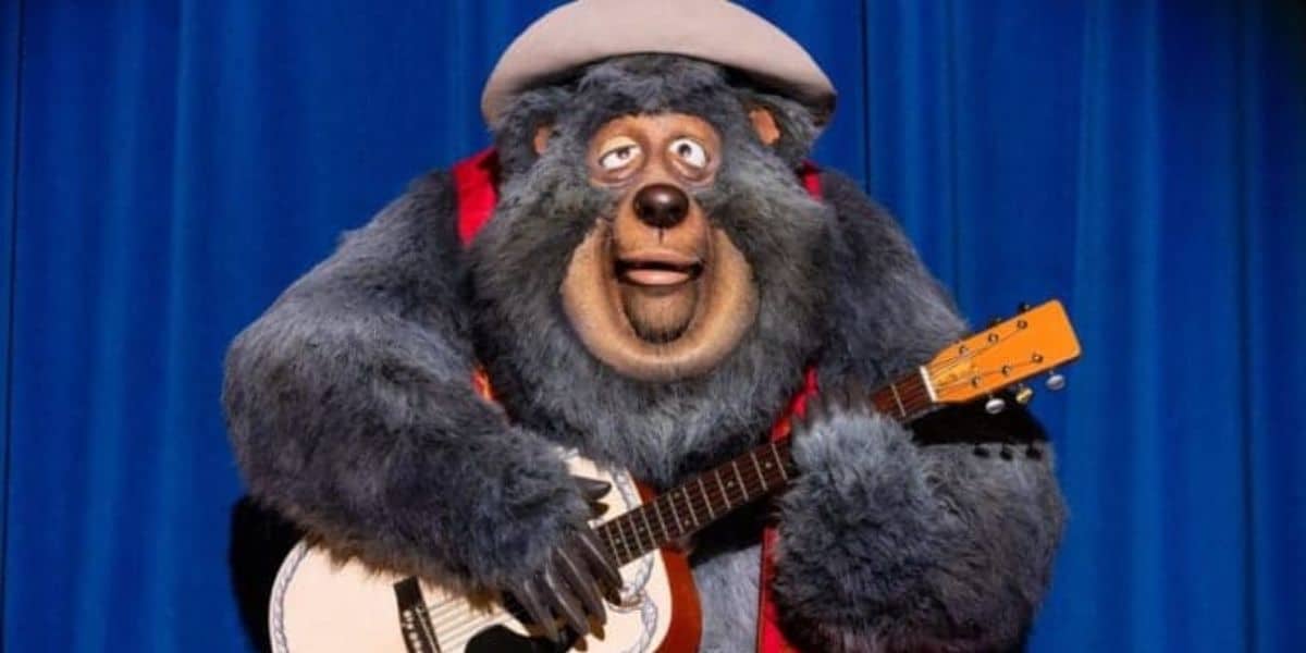 A large anthropomorphic bear wearing a hat and a red vest is joyfully playing a guitar on a stage with a blue curtain backdrop. The bear appears to be part of an animated or animatronic performance.