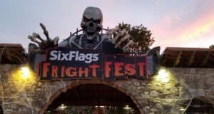 Entrance to Six Flags Fright Fest, featuring a large skeleton with red eyes and outstretched hands above a stone archway. The sky is tinged with pink and orange hues, adding to the eerie atmosphere.