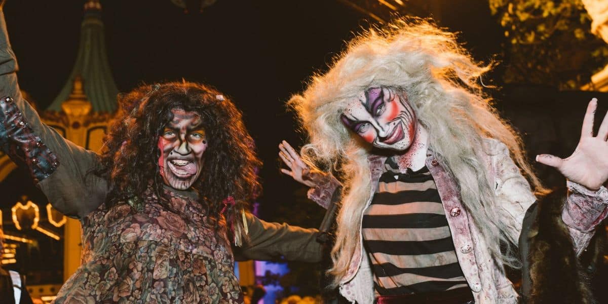 Two people dressed in elaborate Halloween costumes and makeup pose outdoors at night, with a spooky and fun atmosphere. One wears dark curly hair and the other has wild, light-colored hair. Both have exaggerated facial makeup.