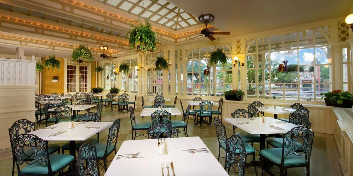 Bright, airy restaurant with large windows and hanging plants offers the best dining at Magic Kingdom Park. Metal lattice chairs surround white tables set with silverware. Warm lighting and intricate ceiling details create a cozy ambiance.