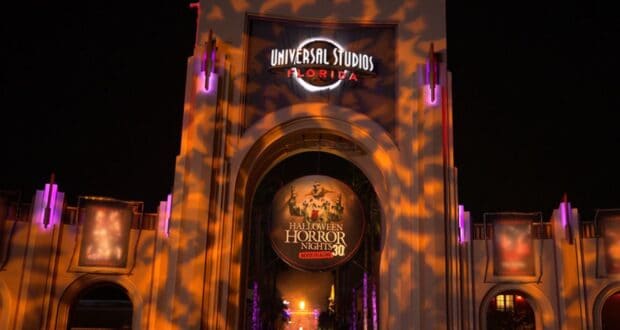 Illuminated entrance of Universal Studios Florida decorated for Halloween Horror Nights 30. The archway displays spooky light patterns and a large circular sign with event details. It is nighttime, enhancing the eerie atmosphere.