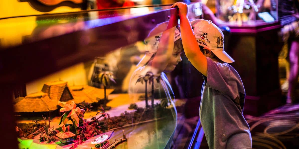 A young child wearing a white cap gazes intently into a display case, their reflection mingling with the hidden gems of a tropical scene. The miniature model showcases small huts and palm trees, all surrounded by bright, colorful lighting worthy of Walt Disney World's Hollywood Studios magic.