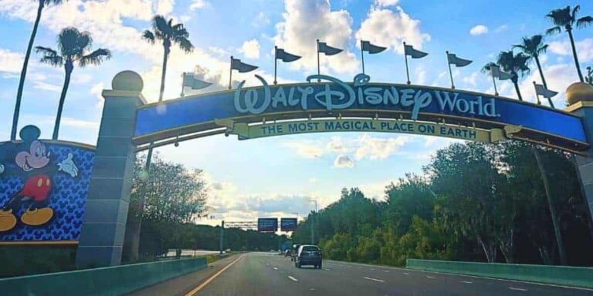 Entrance sign to Walt Disney World with the text "The Most Magical Place on Earth" displayed prominently. A Mickey Mouse image is on the left side of the sign. The road leads past the entrance with trees on both sides and blue sky with clouds overhead.