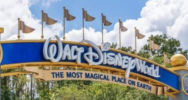 Entrance sign to Walt Disney World with a blue and gold design, featuring the phrase "The Most Magical Place on Earth." The sky is partly cloudy, and trees are visible in the background.