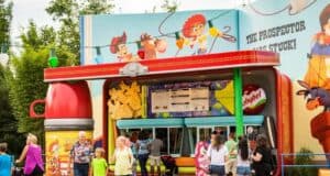 Families and children gather under a clear sky at a colorful, cartoon-themed concession stand with a western motif in Toy Story Land at Walt Disney World. The building showcases playful illustrations of cowboys and prospector characters, capturing the whimsical spirit of the park.