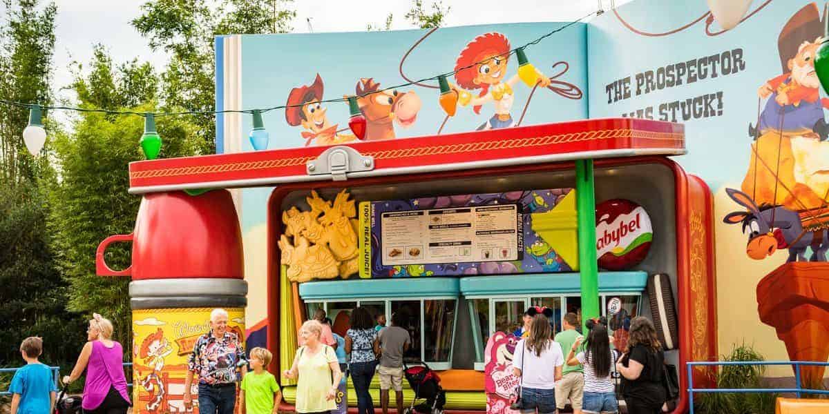 Families and children gather under a clear sky at a colorful, cartoon-themed concession stand with a western motif in Toy Story Land at Walt Disney World. The building showcases playful illustrations of cowboys and prospector characters, capturing the whimsical spirit of the park.
