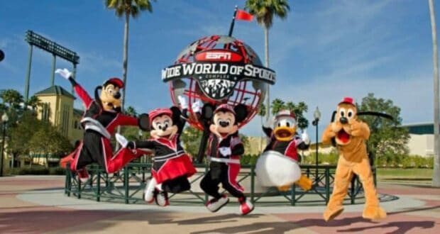 Characters in sports outfits jump in front of the ESPN Wide World of Sports Complex sign. The scene is lively, with palm trees and a clear sky in the background.