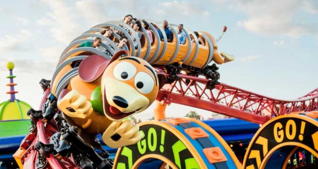 A colorful roller coaster at Disney World, shaped like a springy dog with a cartoonish face, is full of excited riders. The track features bright, playful designs, including loops marked "GO!" against a sunny sky.