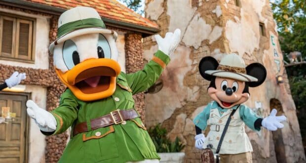 Two Disney characters dressed as explorers stand cheerfully outside a rustic building. The character on the left wears a green safari outfit, and the one on the right wears a beige explorer outfit with a hat. Trees and a wooden structure are in the background.