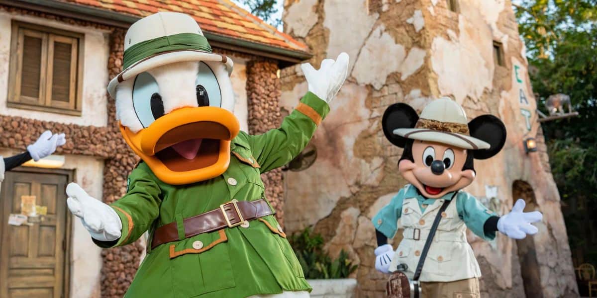 Two Disney characters dressed as explorers stand cheerfully outside a rustic building. The character on the left wears a green safari outfit, and the one on the right wears a beige explorer outfit with a hat. Trees and a wooden structure are in the background.