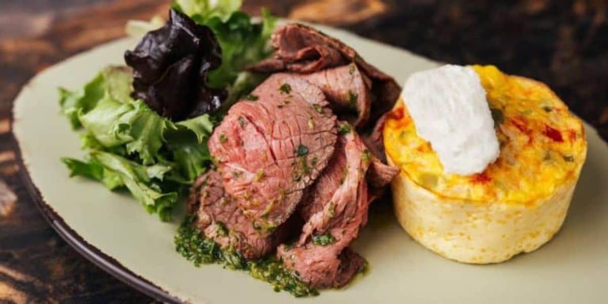 A plate with slices of roast beef in herb sauce, a serving of leafy greens, and a small frittata topped with a dollop of cream.