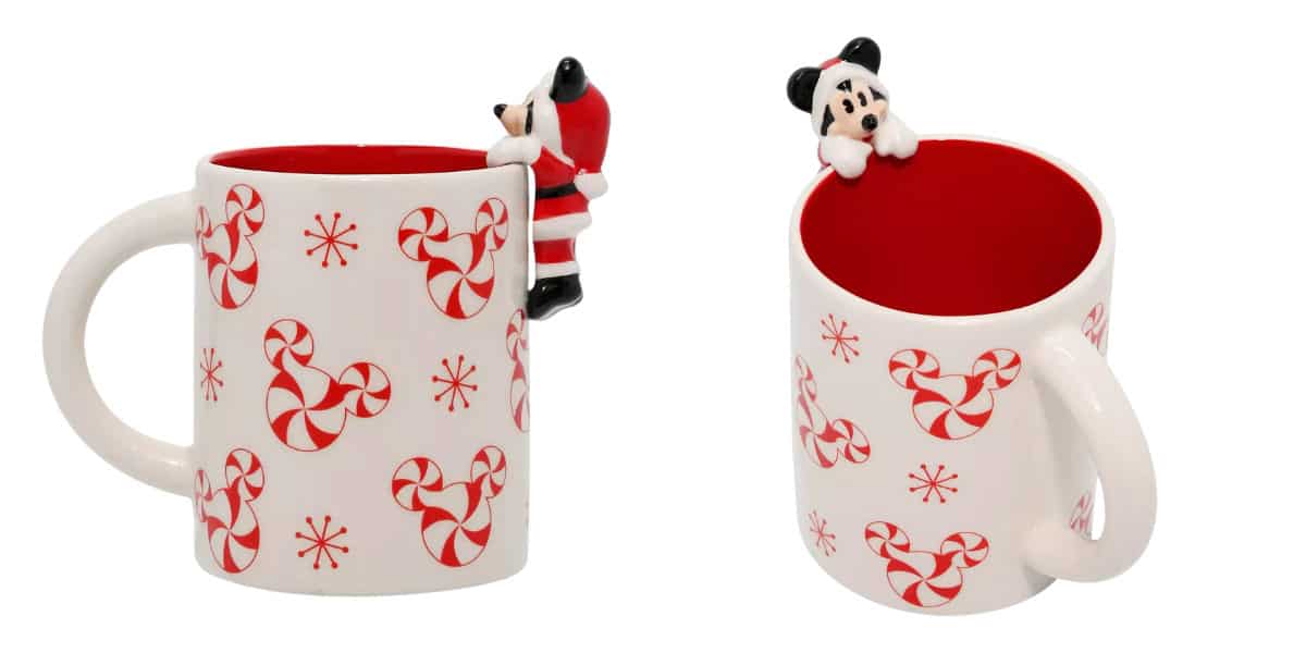 Two white mugs with red interiors are decorated with peppermint swirl designs and snowflakes. Each has a small figurine of a dog wearing a Santa hat climbing on the rim.
