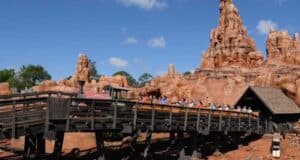 A roller coaster train is navigating a wooden track through a rugged, red rock canyon landscape on a sunny day. The ride has adventurous and western-themed scenery with clear blue skies above.