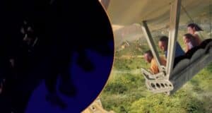 People are on a ride, simulating a flight over a green landscape. They're seated in what appears to be an amusement park ride, with a partially darkened foreground. The image gives a sense of excitement and adventure.