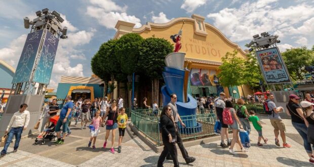 A bustling scene at Walt Disney Studios with a large crowd of people, some pushing strollers. The main building features Disney-themed decorations. Bright, sunny weather with a partly cloudy sky and lush greenery surrounds the area.