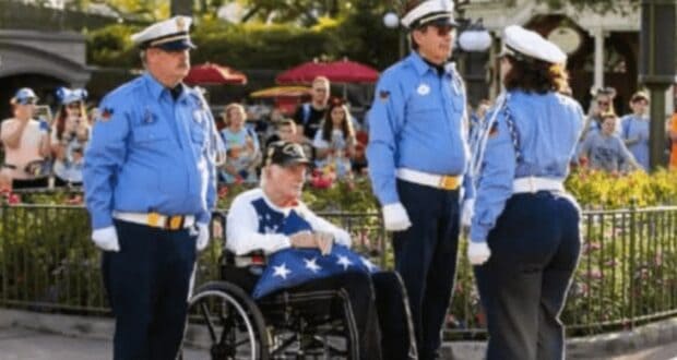 A military veteran in a wheelchair holds a folded American flag, surrounded by three uniformed security personnel. They are in front of a crowd, with greenery and a building in the background.
