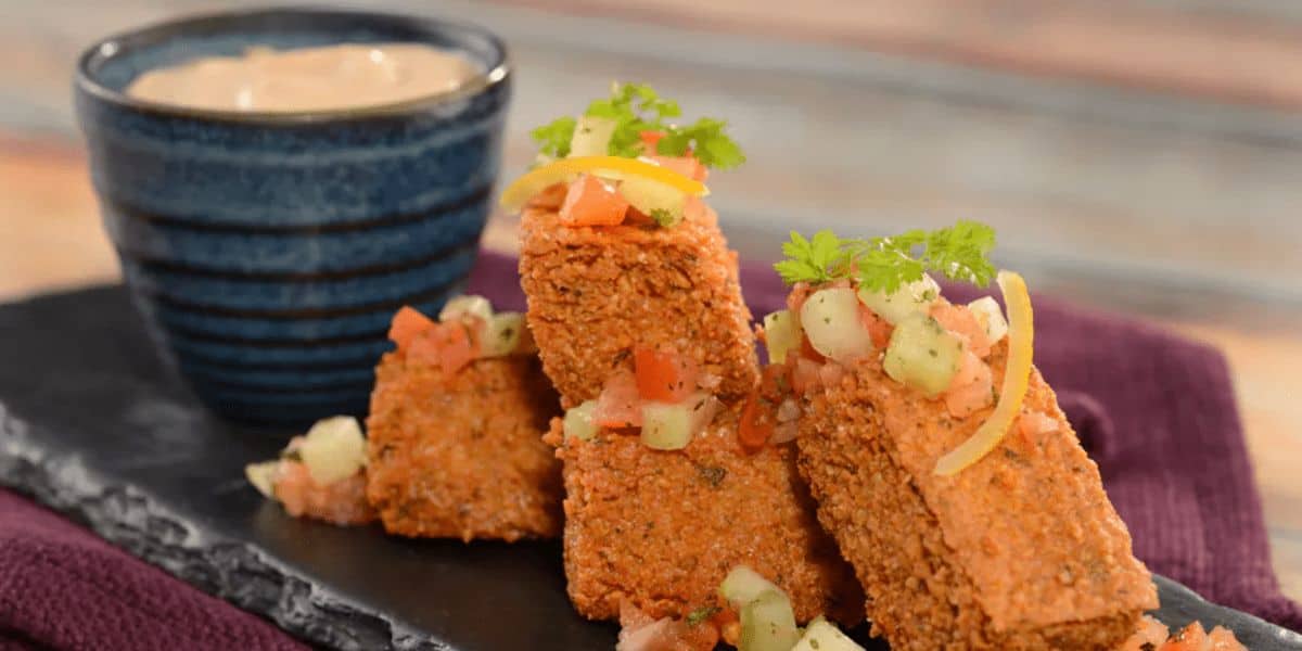 A plate of crispy falafel from Spice Road Table is garnished with diced cucumber, tomatoes, and herbs, arranged in a stack. A small blue bowl of dipping sauce rests beside them on a dark serving board, all elegantly displayed on a purple cloth.