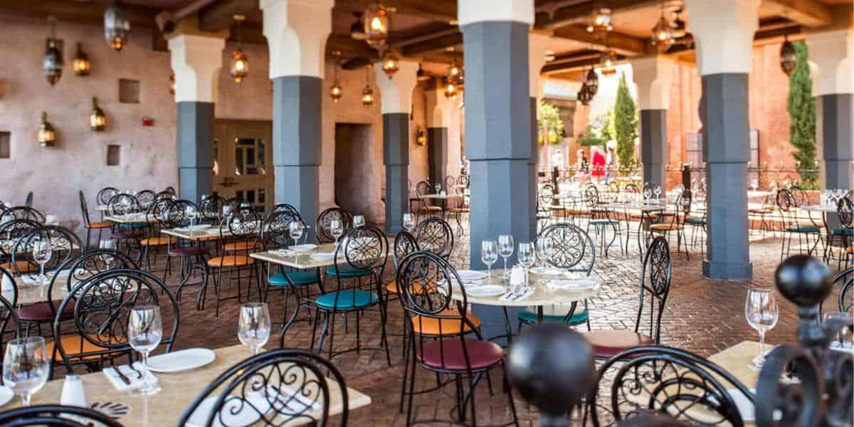 Spice Road Table features an outdoor dining area with elegant wrought-iron chairs adorned with multicolored cushions and stone-tiled floors. Tables are set with white tablecloths, wine glasses, and cutlery. Lanterns hang from the ceiling, and arches add architectural interest.