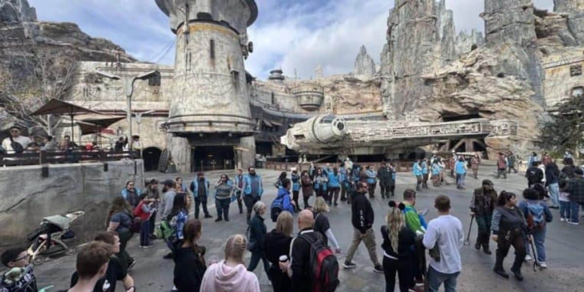 A bustling theme park scene with people gathered around a large, detailed spacecraft and rugged, rocky structures. The sky is partly cloudy, enhancing the adventurous atmosphere of the location.