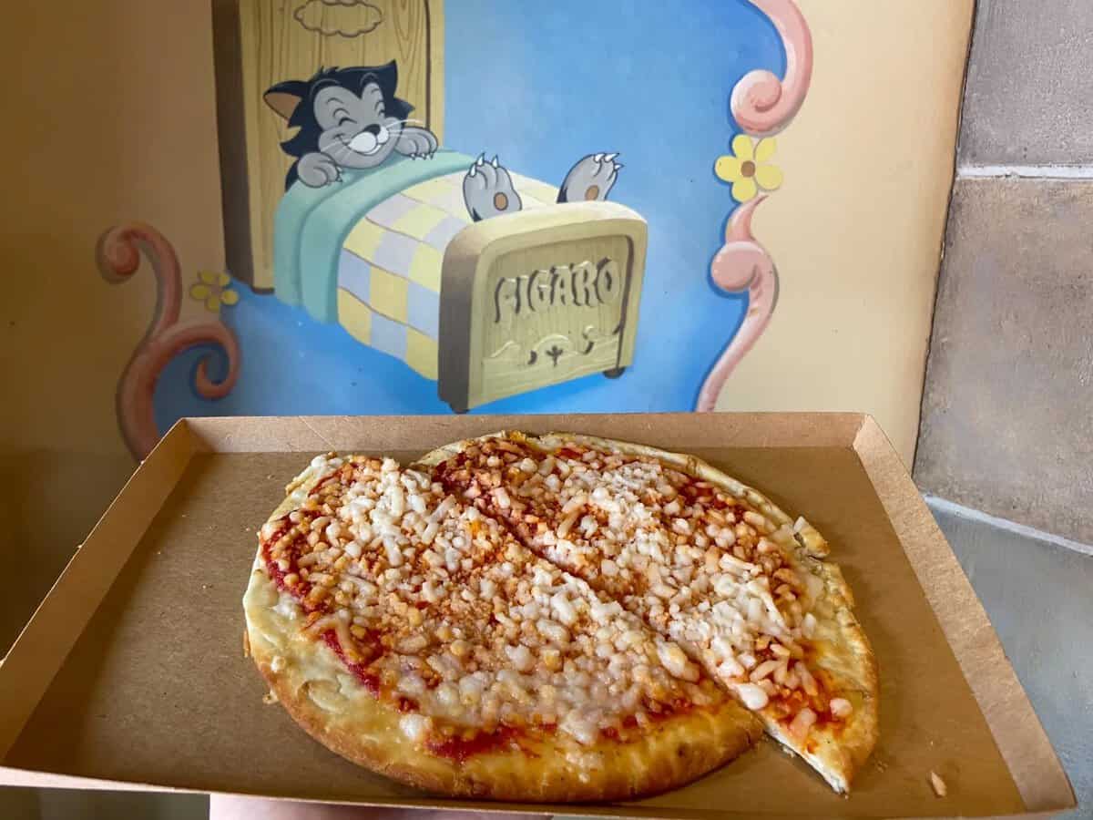 A cheese pizza with a crispy crust is on a cardboard tray. In the background, there's a mural of a cat on a bed labeled "FIGARO," with mice peeking over the edge.