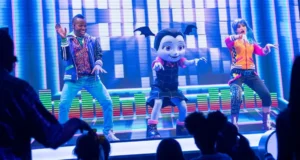 A vibrant stage performance featuring a large, animated vampire bat character in the center flanked by two energetic dancers. The background is colorful with digital graphics, and the audience, mostly children, watches excitedly.
