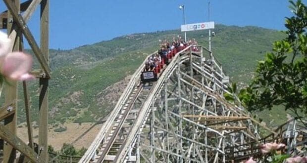 A roller coaster car filled with passengers descends a wooden track under a clear blue sky. The track is surrounded by greenery and a mountain landscape in the background. Pink flowers are visible in the foreground.
