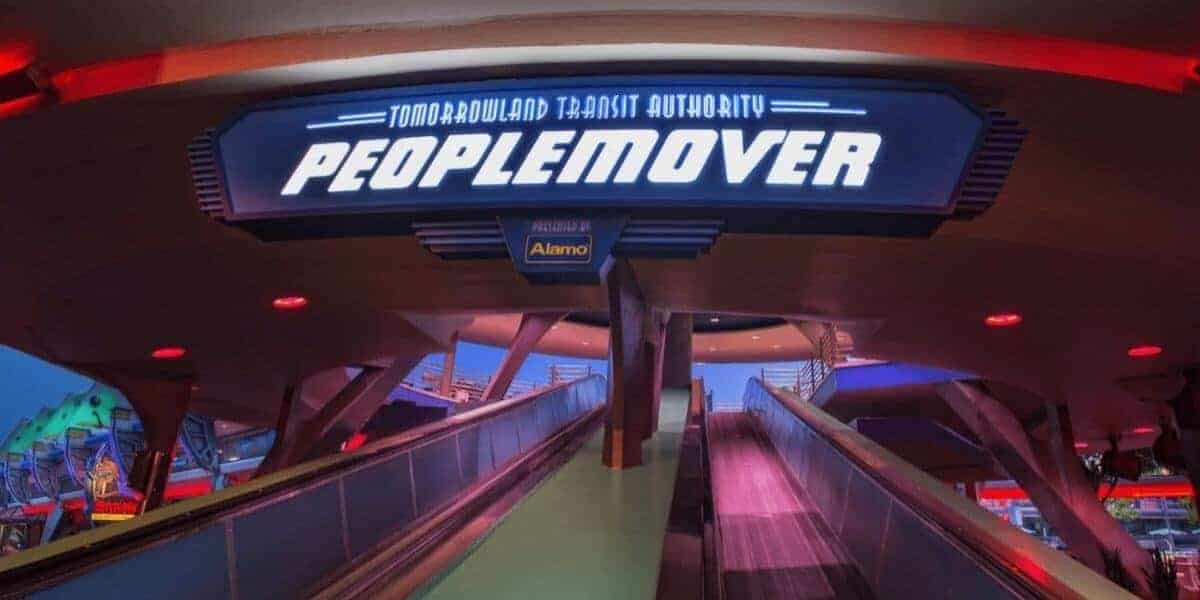 Sign for the Tomorrowland Transit Authority PeopleMover at an amusement park, above an ascending walkway. The area is illuminated with colorful lights, creating a futuristic atmosphere.
