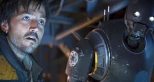 A man with messy hair and a scruffy beard stands next to a tall, black droid with glowing eyes. Both appear tense and are lit by blue and yellow light inside a metallic, industrial setting.
