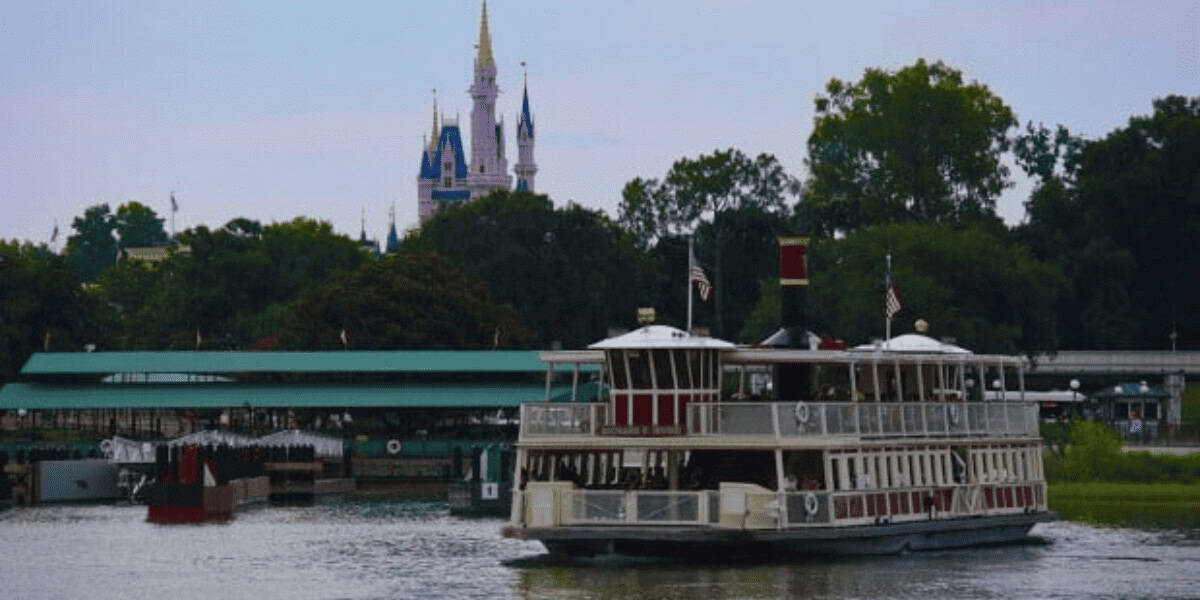 A large white and red ferry boat crosses a calm body of water, with a green-roofed dock and a fairytale castle with blue spires visible in the background among trees.