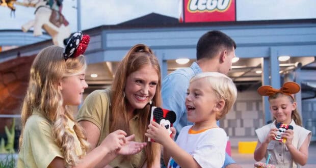 A smiling woman and two children hold Mickey Mouse-themed LEGO creations outside a LEGO store at Disney Springs. In the background, a man and girl wearing mouse ears enjoy more Disney Springs activities, building with colorful LEGO bricks.