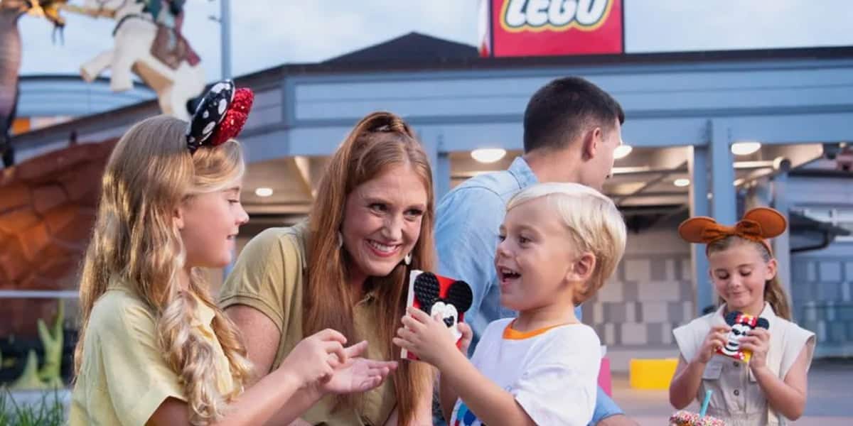 A smiling woman and two children hold Mickey Mouse-themed LEGO creations outside a LEGO store at Disney Springs. In the background, a man and girl wearing mouse ears enjoy more Disney Springs activities, building with colorful LEGO bricks.