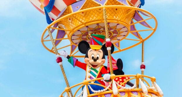 Mickey Mouse, dressed in a colorful festive outfit, stands on a vibrant, elaborate parade float designed like a hot air balloon basket, waving against a clear blue sky.