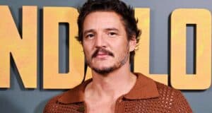 A man with short curly hair, a mustache, and beard stands in front of a wall with large yellow letters. He is wearing a brown, open-knit shirt and looking at the camera with a neutral expression.