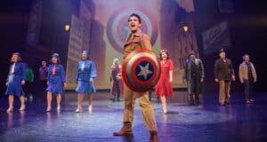 Group of performers in retro costumes sing on stage. One holds a large star-emblazoned shield. A cityscape backdrop adds depth to the scene, with vintage signage visible. The atmosphere is lively and theatrical.