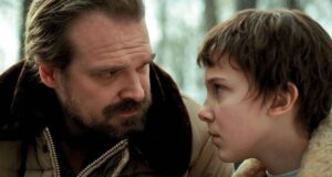 A bearded man in a brown jacket looks seriously at a young person with short hair and a fur-collared coat; both appear to be outdoors in a tense moment.