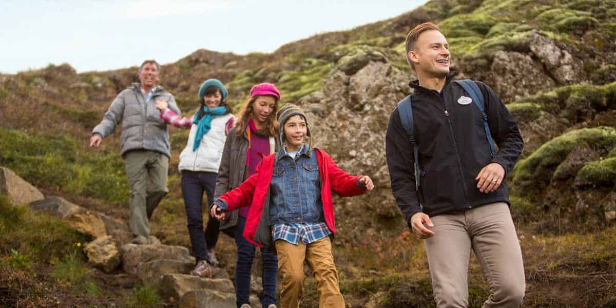 Five people, including adults and children, are smiling as they hike down a rocky, grassy hillside. They appear to be enjoying the outdoors together.